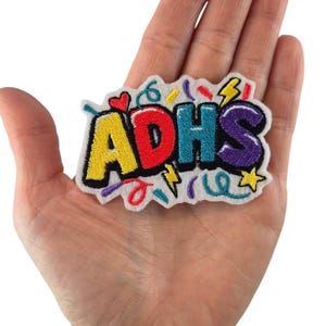 Könnte beinhalten: Ein farbenfroher, gestickter Aufnäher mit den Buchstaben "ADHS" in Gelb, Rot, Türkis und Lila. Der Aufnäher hat einen weißen Hintergrund mit Konfetti und einem Herzmotiv. Der Aufnäher wird in einer Hand gehalten.