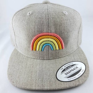 Könnte beinhalten: Eine graue Baseballkappe mit einem aufgestickten Regenbogen auf der Vorderseite. Die Kappe hat ein schwarz-weißes Etikett mit der Aufschrift "The Authentic Yupoong Snapback Yupoong Since 1974".