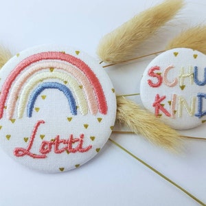 Könnte beinhalten: Zwei gestickte Stoffabzeichen. Eines zeigt einen Regenbogen mit dem Namen "Lotti" darunter. Das andere Abzeichen sagt "Schulkind" in bunten Buchstaben.