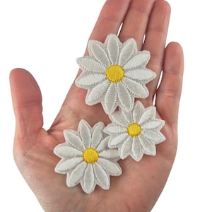 Op de afbeelding: Drie witte madeliefjes bloem applicaties met gele centra worden in een hand getoond. De geborduurde applicaties hebben gedetailleerde bloemblaadjes en zijn ideaal voor knutselen of het verfraaien van kleding en accessoires.