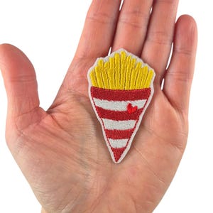 Könnte beinhalten: Gestickter Aufnäher mit einer Pommes-Tüte. Die Oberseite der Tüte hat gelbe Pommes, und die Tüte ist rot-weiß gestreift mit einem kleinen roten Herz. Der Aufnäher wird in einer Hand gehalten.