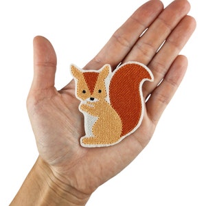 May include: A brown and orange embroidered squirrel patch with white accents. The patch is on a hand.