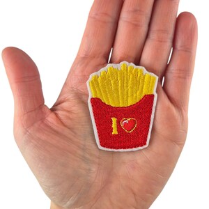 May include: Embroidered patch featuring a red container filled with yellow french fries. The patch includes the text "I <3" with a heart symbol. The patch is held in a person's hand.