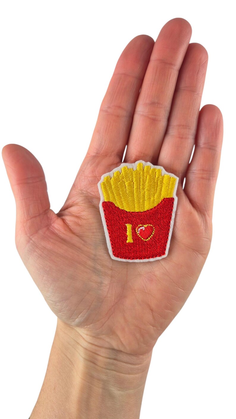 May include: Embroidered patch featuring a red container filled with yellow french fries. The patch includes the text "I <3" with a heart symbol. The patch is held in a person's hand.