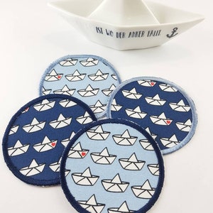 May include: Four round coasters with a navy blue trim and a paper boat pattern in light and dark blue. A white ceramic boat-shaped dish with the text "IST WO DER ANKER FÄLLT" is in the background.