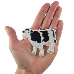 Könnte beinhalten: Ein gestickter Bügel-Patch mit einer schwarz-weißen Kuh mit rosa Akzenten. Der Patch befindet sich auf einer Hand zur Größenreferenz.