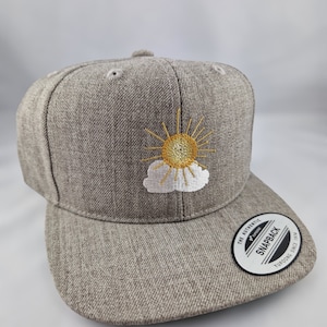 Könnte beinhalten: Eine graue, bestickte Baseballkappe mit einem gelben Sonnen- und einem weißen Wolkenmotiv. Die Kappe hat ein schwarz-weißes Etikett mit der Aufschrift "The Authentic Yupoong Snapback Yupoong Since 1974".