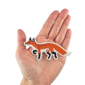 May include: An orange embroidered fox patch with black details. The patch is on a hand, showing its size.