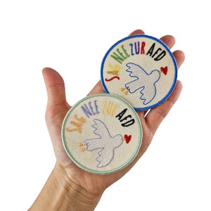 Può includere: Due toppe rotonde ricamate con una colomba bianca e il testo "Sag Nee zur AfD" in colori arcobaleno. Le toppe sono su una mano.