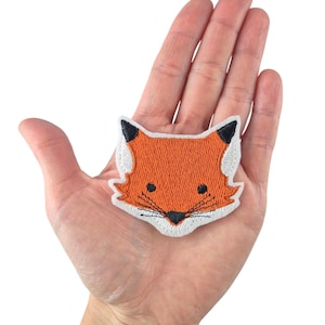 Könnte beinhalten: Ein kleiner gestickter Fuchskopf-Patch in Orange mit weißen und schwarzen Details. Der Patch befindet sich auf einer Hand.