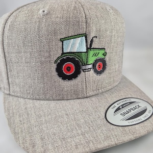 Könnte beinhalten: Eine graumelierte Snapback-Cap mit einem gestickten grünen Traktor, schwarzen Reifen und roten Radmitten. Die Cap hat einen runden schwarz-weißen Aufkleber auf dem flachen Schirm mit der Aufschrift The Authentic Yupoong Snapback Since 1974.