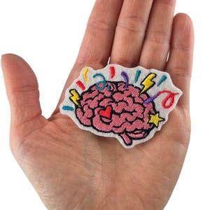Pode incluir: Patch bordado com um cérebro rosa com um coração vermelho, rodeado por elementos festivos coloridos. O patch, sobre um fundo branco, tem detalhes em amarelo, vermelho e azul. O patch é segurado em uma mão.