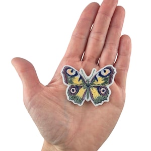 Könnte beinhalten: Ein gestickter Schmetterlings-Patch in Gelb und Grün mit schwarzen und weißen Augen. Der Patch befindet sich auf einem weißen Hintergrund.