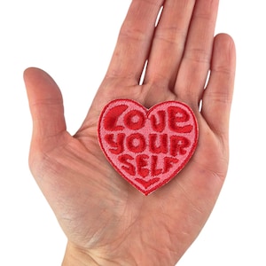 Peut inclure: Un écusson brodé en forme de cœur, dans des tons de rouge et de rose, avec les mots "LOVE YOUR SELF" brodés en gras. L'écusson est tenu dans une main ouverte sur fond blanc. L'écusson mesure environ 5 cm de large.