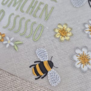 May include: A white background with green text that reads "Bienchen & Bl&uuml;mchen pssssso". There are yellow and white flowers and a black and yellow bee with gray wings.