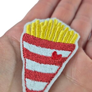 May include: Embroidered patch featuring a stylized design of a container of french fries. The container is red and white striped, with a small red heart. The fries are yellow.