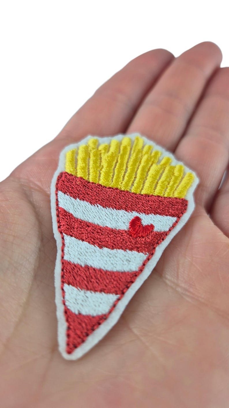 May include: Embroidered patch featuring a stylized design of a container of french fries. The container is red and white striped, with a small red heart. The fries are yellow.