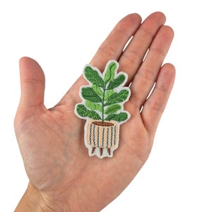 Könnte beinhalten: Eine grüne gestickte Pflanze in einem braunen und weißen gestreiften Topf mit braunen Beinen. Die Pflanze befindet sich auf einem weißen Hintergrund.