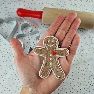 Könnte beinhalten: Ein Lebkuchenmann-Ausstecher mit einem lächelnden Gesicht und einem roten Herzen auf der Brust. Der Ausstecher ist aus Metall und hat einen weißen Umriss.