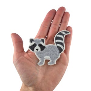 Könnte beinhalten: Gestickter Aufnäher mit einem grauen Waschbären mit schwarzen Markierungen und einem gestreiften Schwanz. Der Aufnäher hat einen weißen Rand und ist zum Aufbügeln oder Aufnähen auf Stoff vorgesehen. Ideal zum Dekorieren von Kleidung oder Accessoires.