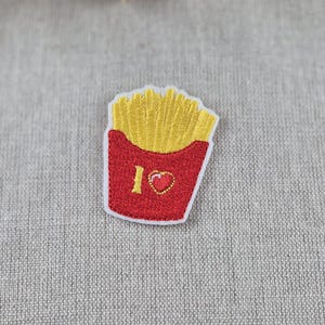 May include: Embroidered patch featuring a red container of golden french fries. The patch includes the text "I ❤️" with a heart symbol. The patch is on a gray fabric background.