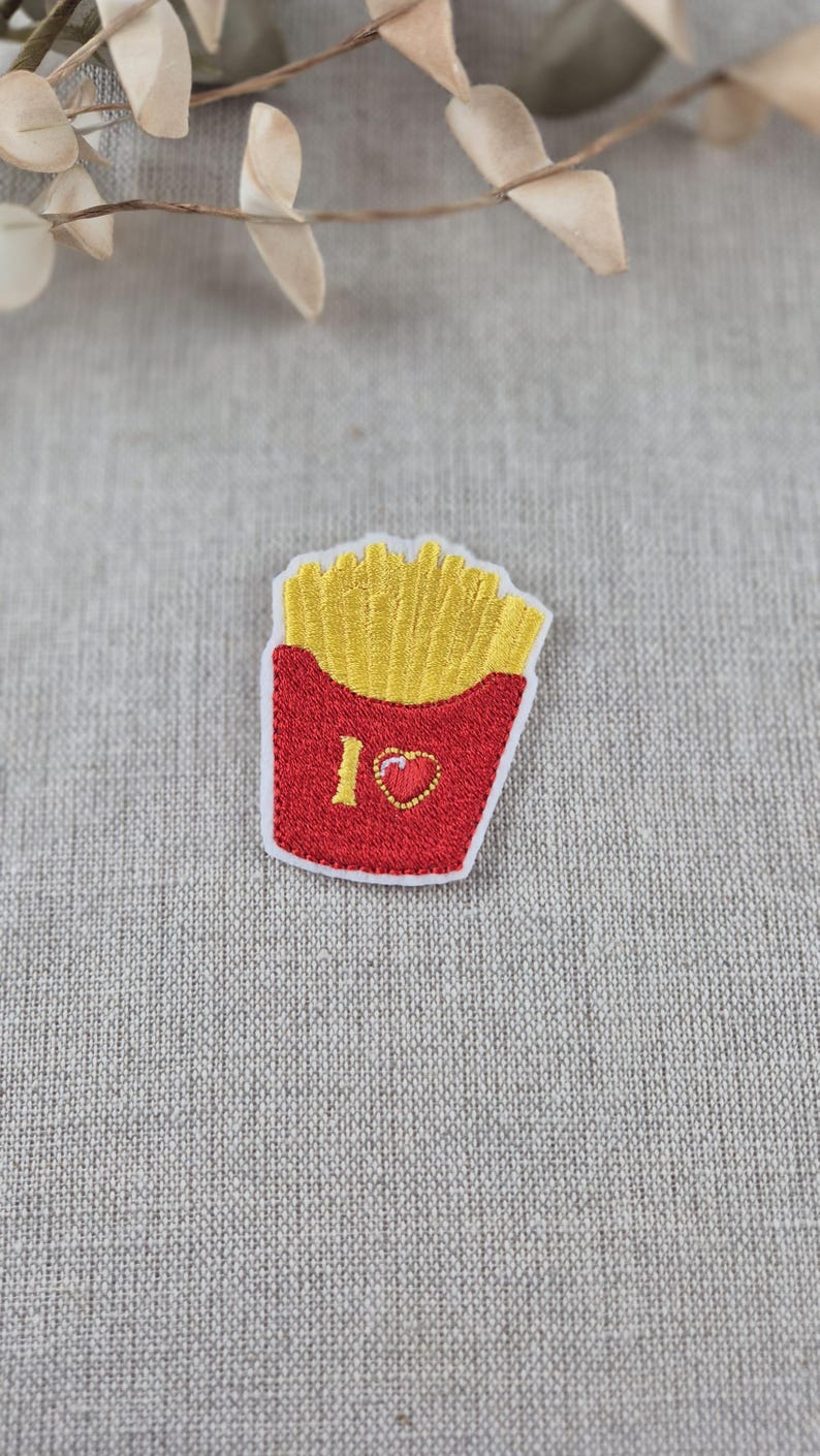 May include: Embroidered patch featuring a red container of golden french fries. The patch includes the text "I ❤️" with a heart symbol. The patch is on a gray fabric background.