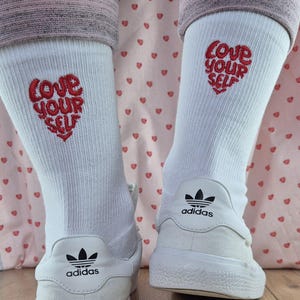 Könnte beinhalten: Weiße Socken mit einem roten, gestickten Herz und dem Schriftzug "Love Your Self". Die Socken werden mit weißen Adidas-Sneakern getragen. Das Adidas-Logo ist auf der Rückseite der Sneaker zu sehen. Der Hintergrund ist ein rosa Stoff mit kleinen roten Herzen.