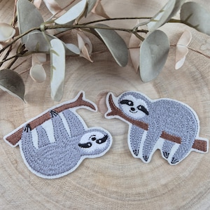 May include: Two embroidered patches featuring sloths. One patch shows a sloth hanging from a brown branch, and the other shows a sloth sitting on a branch. Both sloths are gray with black and white details. The patches are on a wooden surface.