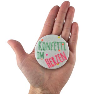Könnte beinhalten: Ein weißer runder Knopf mit grüner und pinkfarbener Stickerei, auf dem "Konfetti im Herzen" steht, mit bunten Konfettipunkten um den Text herum.