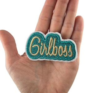 Peut inclure: Un écusson brodé vert d'eau et doré avec le mot "Girlboss" en écriture cursive. L'écusson a une bordure blanche et est tenu dans une main. L'arrière-plan est blanc. Un article décoratif.