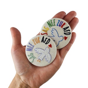 Könnte beinhalten: Zwei weiße, bestickte Stoffknöpfe mit einem regenbogenfarbenen Text, der "Say No To Aid" lautet, und einer weißen Taube mit einem roten Herzen.