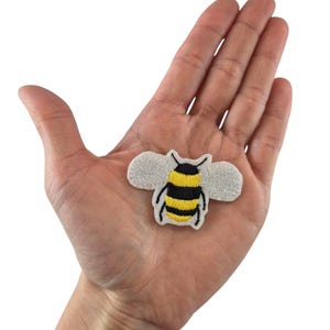 Könnte beinhalten: Ein gestickter Bienen-Patch aus weißem Filz mit schwarzen und gelben Details. Die Biene fliegt mit ausgebreiteten Flügeln.