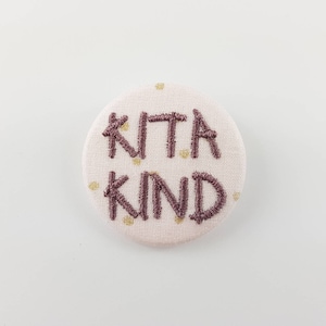 Op de afbeelding: Een witte stoffen knoop met een roze geborduurde boodschap die "Wees aardig" zegt met gouden stippen.