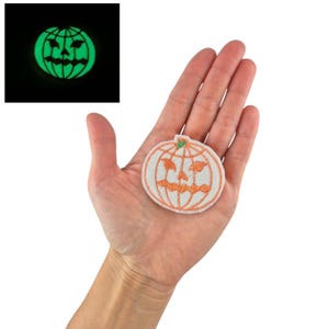 Könnte beinhalten: Ein weißer gestickter Bügelaufkleber mit einer orangefarbenen Kürbis mit einem Jack-o'-Lantern-Gesicht. Der Aufkleber leuchtet im Dunkeln.