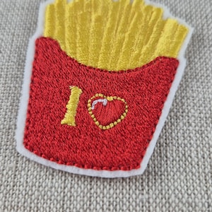 May include: Embroidered patch featuring a red container of golden french fries. The patch includes the text "I ❤️" with a heart symbol. The patch has a white border and is on a textured, neutral background.
