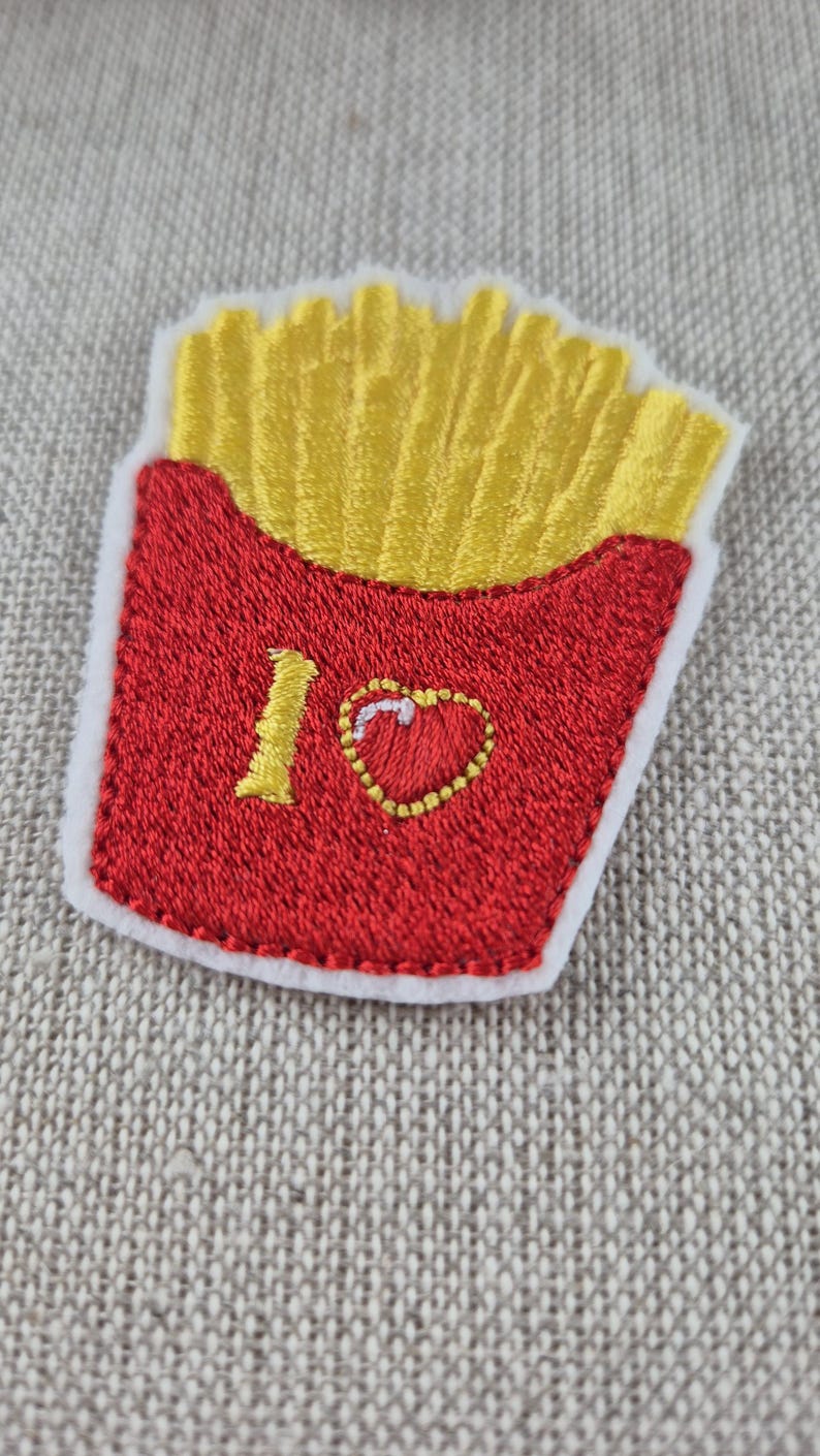 May include: Embroidered patch featuring a red container of golden french fries. The patch includes the text "I ❤️" with a heart symbol. The patch has a white border and is on a textured, neutral background.