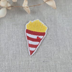 May include: Embroidered patch featuring a stylized cone of french fries. The top portion is yellow, representing the fries, and the bottom is red and white striped with a small red heart. The patch is on a neutral linen background.