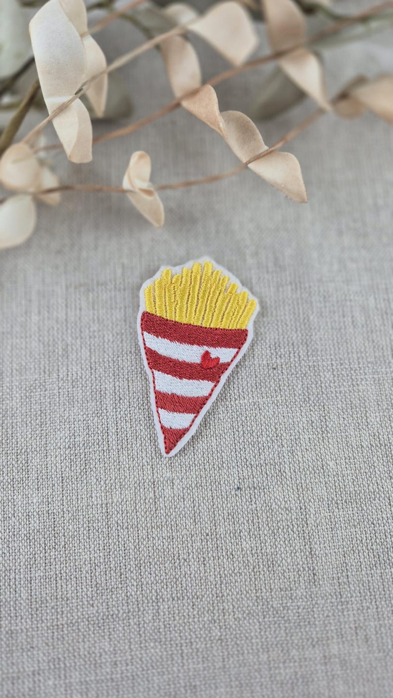 May include: Embroidered patch featuring a stylized cone of french fries. The top portion is yellow, representing the fries, and the bottom is red and white striped with a small red heart. The patch is on a neutral linen background.