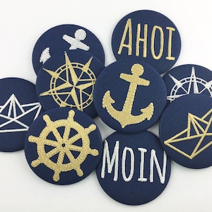 Peut inclure: Neuf boutons ronds en tissu bleu marine avec des broderies dorées et blanches. Les boutons présentent des thèmes nautiques tels qu'une ancre, une boussole, un gouvernail de navire et un bateau en papier. Les textes "AHOI" et "MOIN" sont également brodés sur deux des boutons.