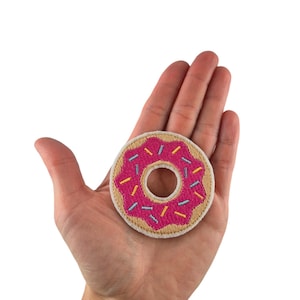 Könnte beinhalten: Ein beigefarbener Stoff-Patch mit einem gestickten rosafarbenen Donut mit bunten Streuseln.