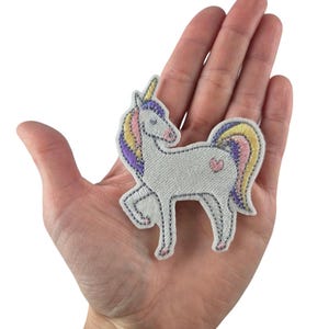 Könnte beinhalten: Ein gestickter Einhorn-Patch in Weiß mit einer Regenbogenmähne und einem Regenbogen-Schwanz. Das Einhorn hat ein pinkfarbenes Herz auf seinem Hinterteil.