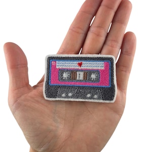 Könnte beinhalten: Gestickter Aufnäher in Form einer Retro-Kassette. Der Aufnäher hat ein rosa und blaues Design mit einem kleinen roten Herz. Die Kassette ist schwarz umrandet und hat einen weißen Hintergrund.