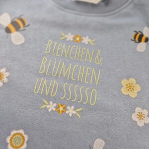 May include: A light blue sweatshirt with embroidered bees, flowers, and the text "Bienchen & Bl&uuml;mchen und sssssso".