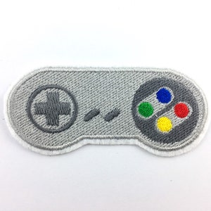Pode incluir: Patch bordado em forma de um controlador de videogame clássico. O patch cinza tem uma borda branca. Possui um direcional, botões coloridos em vermelho, verde, azul e amarelo e dois botões pequenos.
