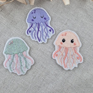 Könnte beinhalten: Drei gestickte Quallen-Patches in Lila, Grün und Pfirsich. Jeder Patch hat einen weißen Rand und detaillierte Nähte mit zarten Tentakeln und Herzakzenten. Die Quallen haben lächelnde oder neutrale Ausdrücke.