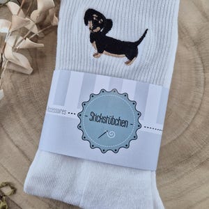 Könnte beinhalten: Weiße Socken mit einer Stickerei eines schwarz-braunen Dackelhundes. Die Socken sind mit einem Papierband mit dem Text "- Stricksfübchen -" in blau-weißem Design umwickelt. Auf einer Holzoberfläche.