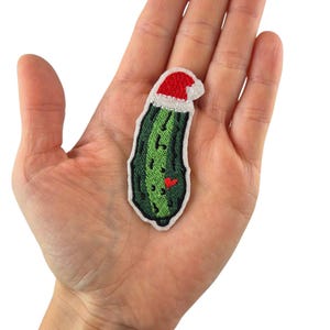 Może przedstawiać: Haftowana naszywka przedstawiająca zielonego ogórka w czerwonej czapce Świętego Mikołaja. Ogórek ma małe czerwone serce i czarne nasiona. Naszywka ma białą obwódkę i jest trzymana w dłoni.