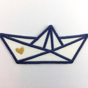 Peut inclure: Un bateau en papier blanc avec un contour bleu et un cœur doré au centre. Le bateau est un design simple avec des bords pliés.