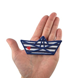 Könnte beinhalten: Ein Aufnäher in Form eines Papierbootes mit marineblauen und weißen Streifen und einem roten Herz. Der Aufnäher ist mit einer marineblauen Kontur bestickt und wird in einer Hand vor weißem Hintergrund gehalten.