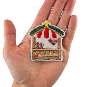 Könnte beinhalten: Gestickter Aufnäher mit einem festlichen Weihnachtsmarktstand. Der Stand hat eine rot-weiß gestreifte Markise, ein grünes Dach und ist mit Lebkuchenmännern und herzförmigen Keksen verziert. Ein gelber Stern krönt den Stand.